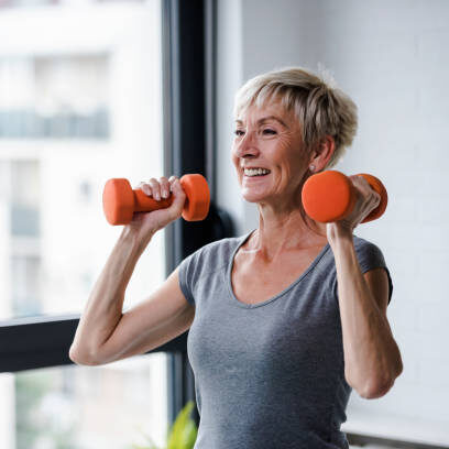 Image of a dietitian holding a stack of papers in hand which are test results. Active good looking elderly woman smiling and holding dumbbells while working out indoors