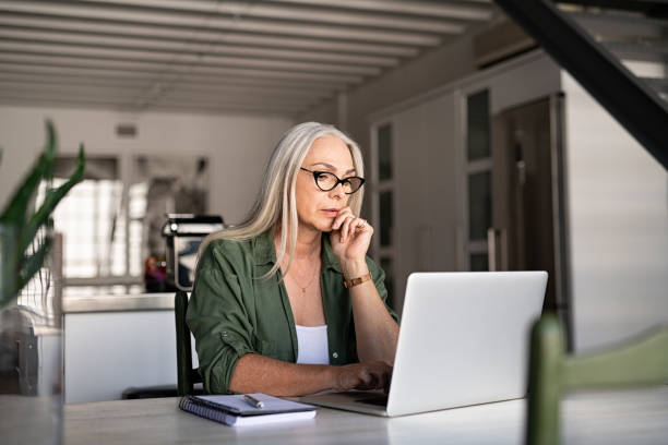 Image of a dietitian holding a stack of papers in hand which are test results. Focused old woman with white hair at home using laptop. Senior stylish entrepreneur with notebook and pen wearing eyeglasses working on computer at home. Serious woman analyzing and managing domestic bills and home finance.