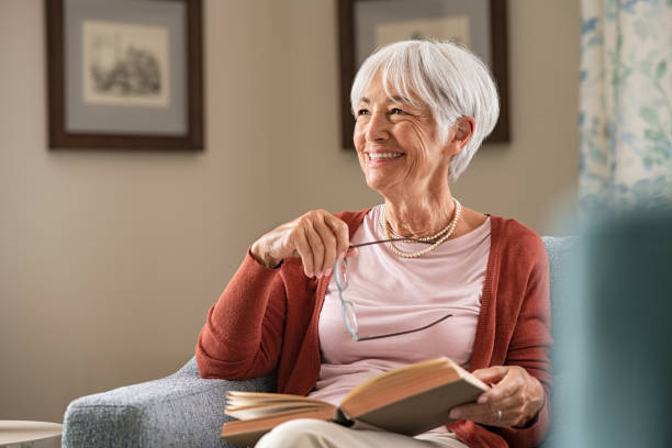 Image of a dietitian holding a stack of papers in hand which are test results. Cheerful senior woman holding book and eyeglasses thinking while relaxing at home. Happy elderly woman reading book at home sitting on couch. Beautiful old teacher takes a break from reading while looking through the window with a big grin.
