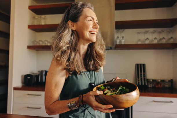 Image of a dietitian holding a stack of papers in hand which are test results. Aging woman smiling happily while holding a buddha bowl in her kitchen. Happy senior woman serving herself a healthy vegan meal at home. Mature woman taking care of her body with a plant-based diet.