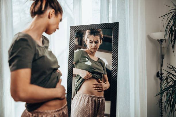 Image of a dietitian holding a stack of papers in hand which are test results. Frown young woman standing in front of a mirror and holding hands on her bloating stomach.