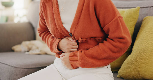 Image of a dietitian holding a stack of papers in hand which are test results. Stomach ache, cramps and hands of woman with abdomen pain due to constipation, menstruation or ibs issue. Sick, home and person suffering and holding belly in a house lounge, couch and living room