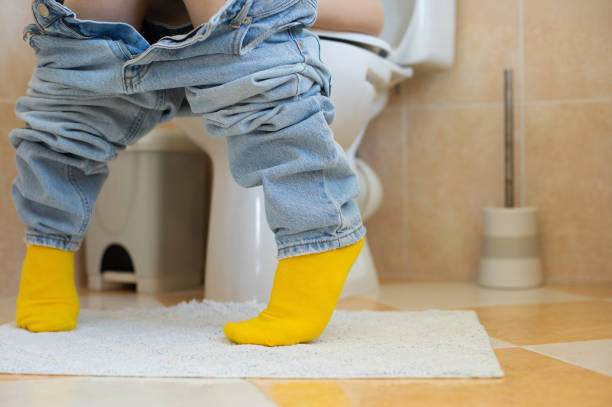 Image of a dietitian holding a stack of papers in hand which are test results. A close-up of a person sitting on a toilet with jeans pulled down and wearing bright yellow socks. The bathroom features a tiled floor and toilet accessories, capturing a relatable, everyday moment.
